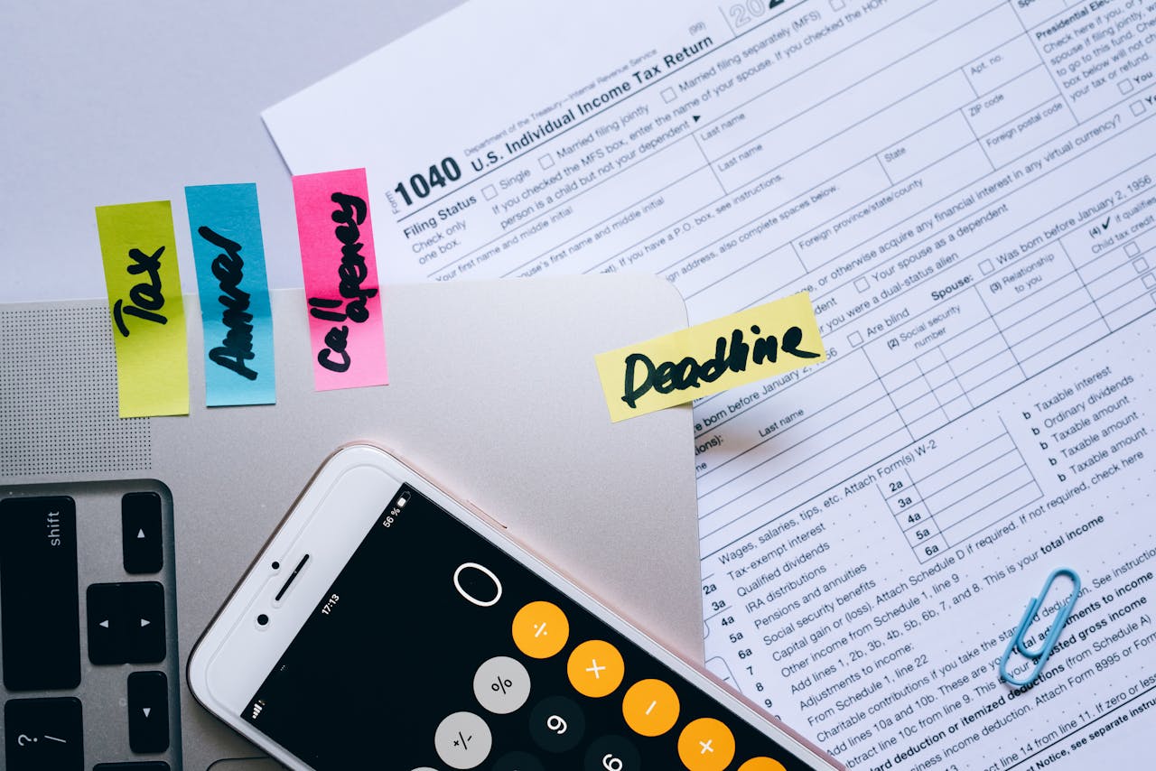 about-us Office desk flat lay showing tax documents, calculator app on smartphone, sticky notes, and paperclips.
